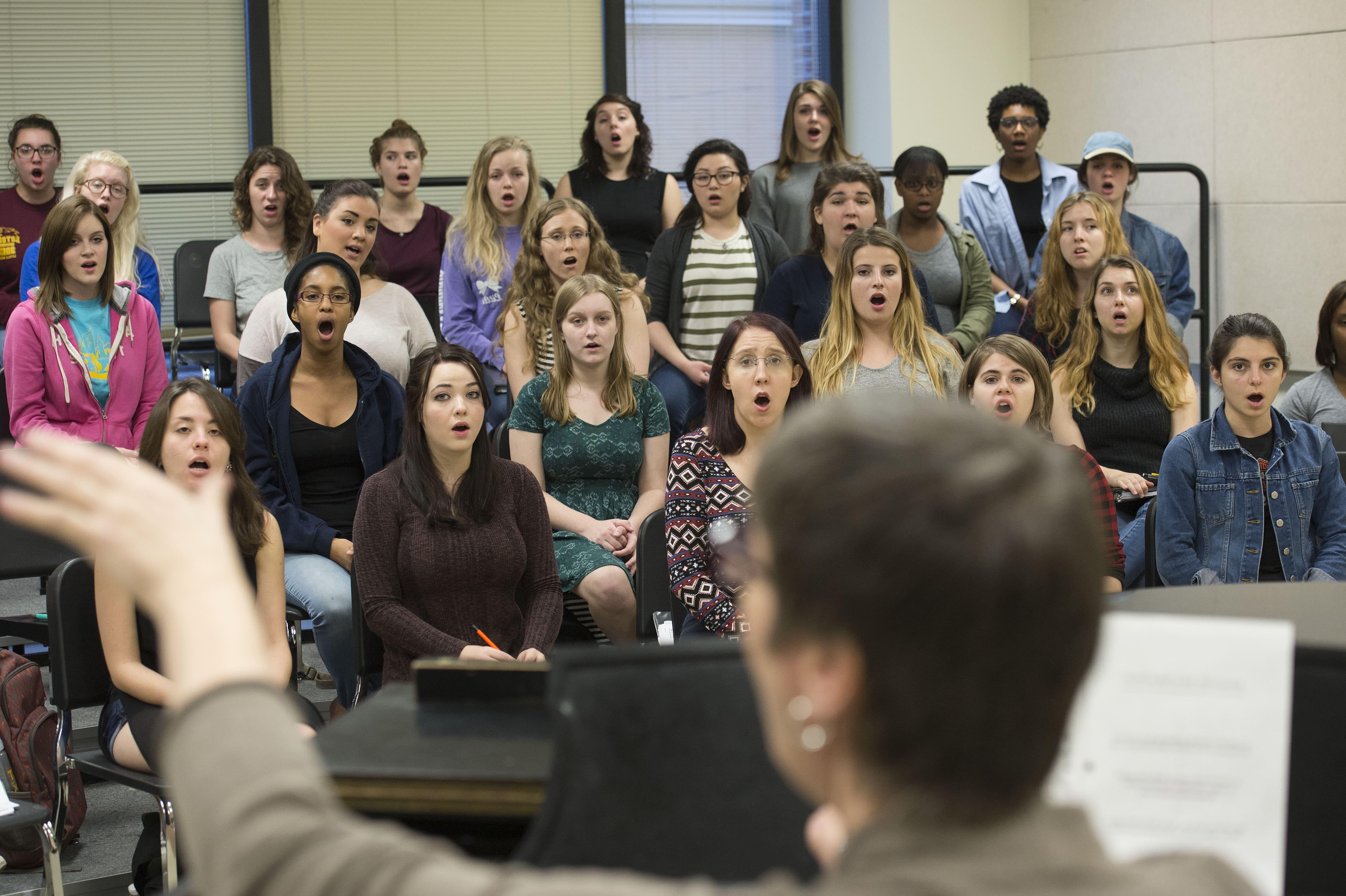 A photo of a choir from behind the shoulder of the conductor, conducting at the piano in a rehearsal setting.